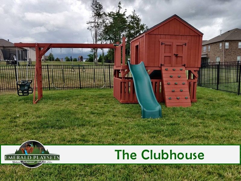 Red wooden clubhouse playset with swing set, climbing wall, and green slide on a grassy lawn.