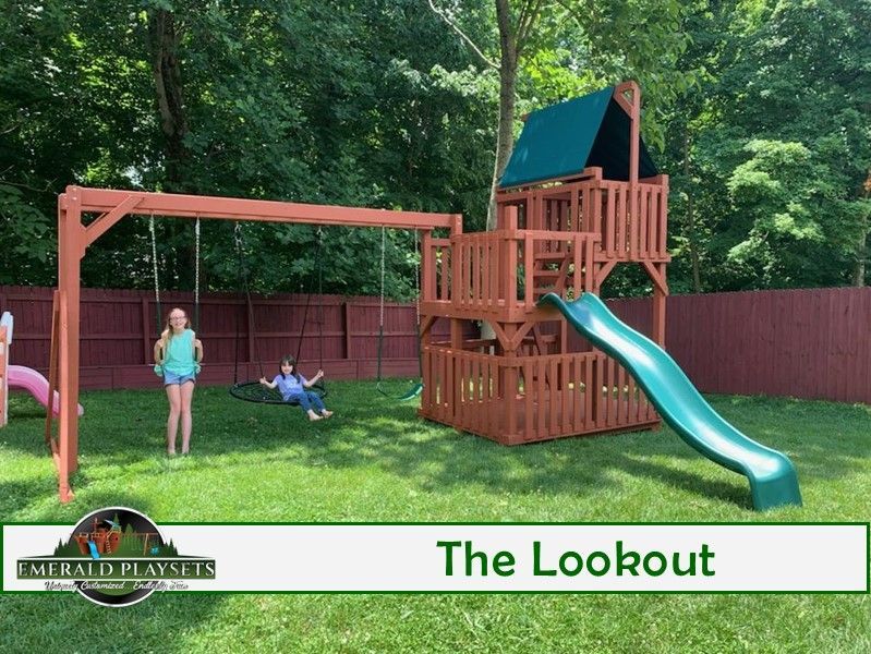 Two children play on a wooden playset in a grassy yard, swing set and slide.