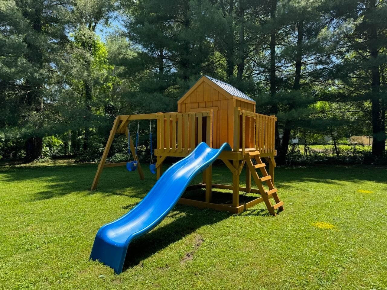 Wooden playset with a blue slide, swings, and a small house on a green lawn.