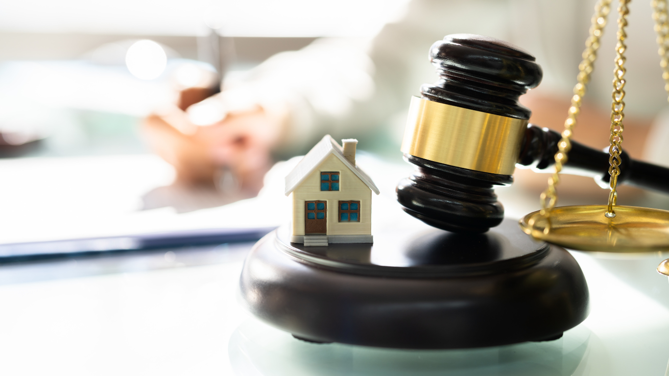 Gavel next to a miniature house, scales, and blurred person; a legal concept.