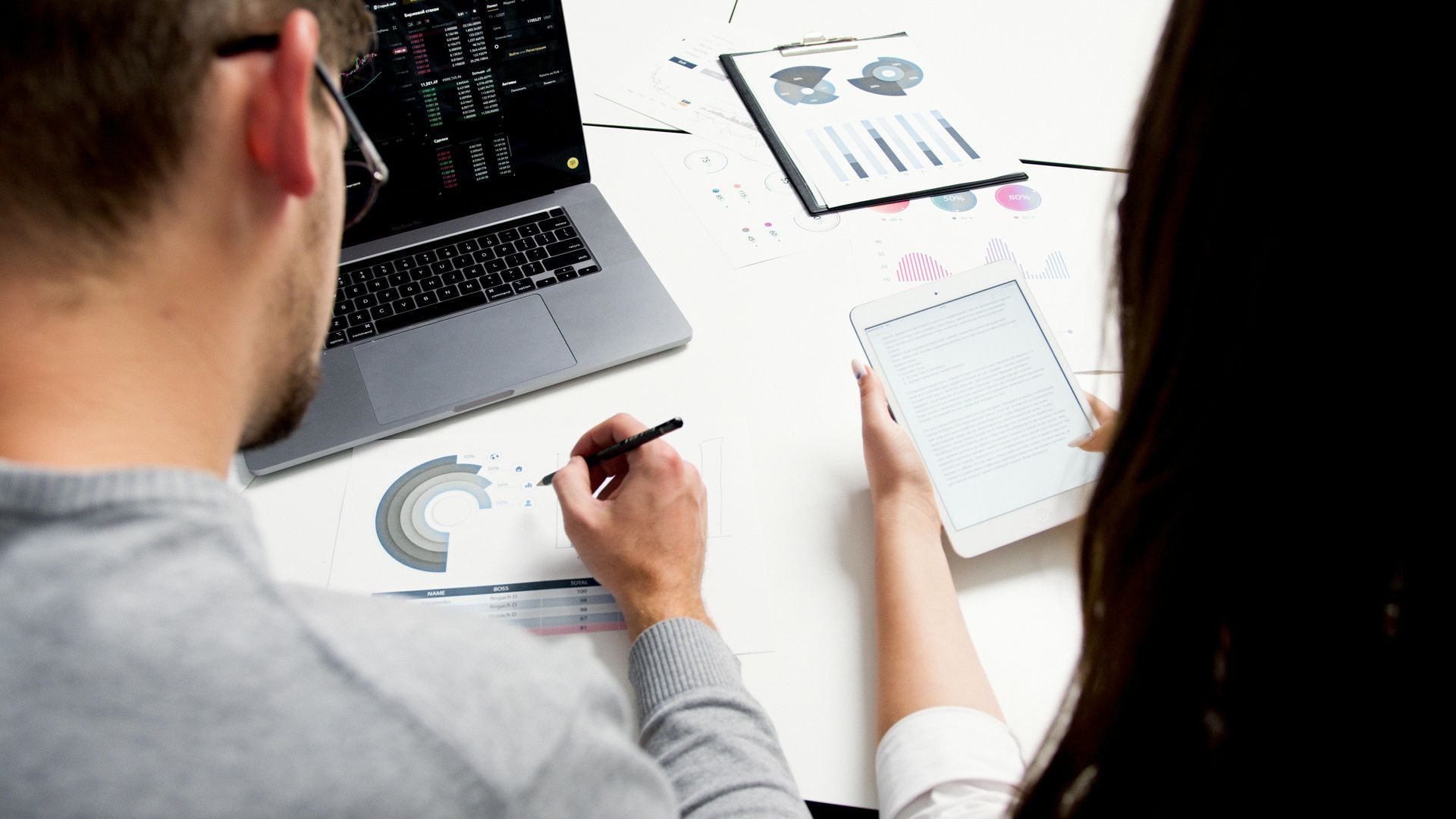Two people working together at a desk with a laptop, tablet, and financial charts.