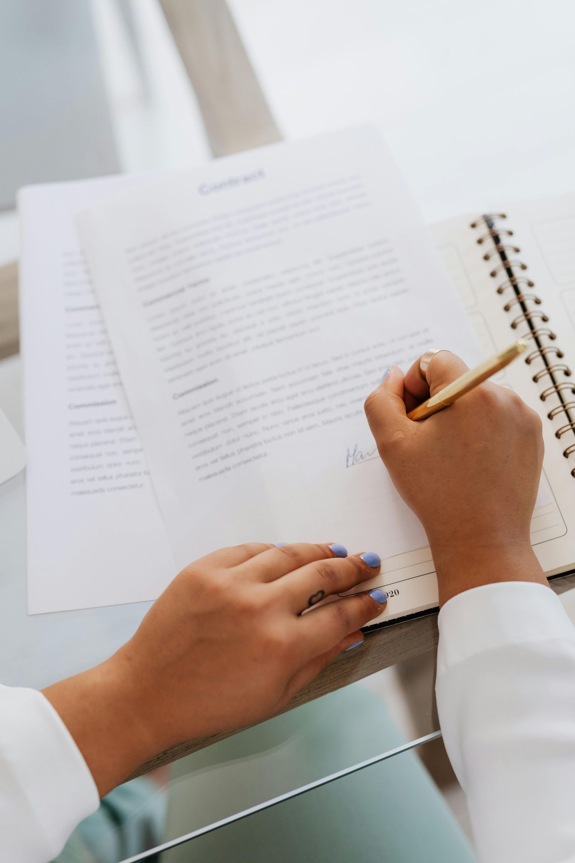Person signing a document with a gold pen, beside an open spiral notebook on a white surface.