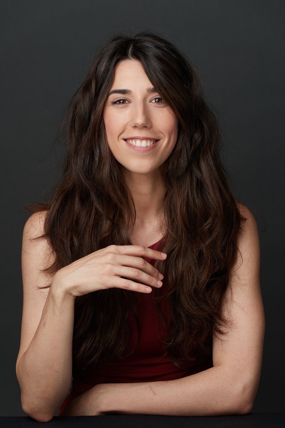 Una mujer con cabello largo está sentada en una mesa con los brazos cruzados y sonriendo.