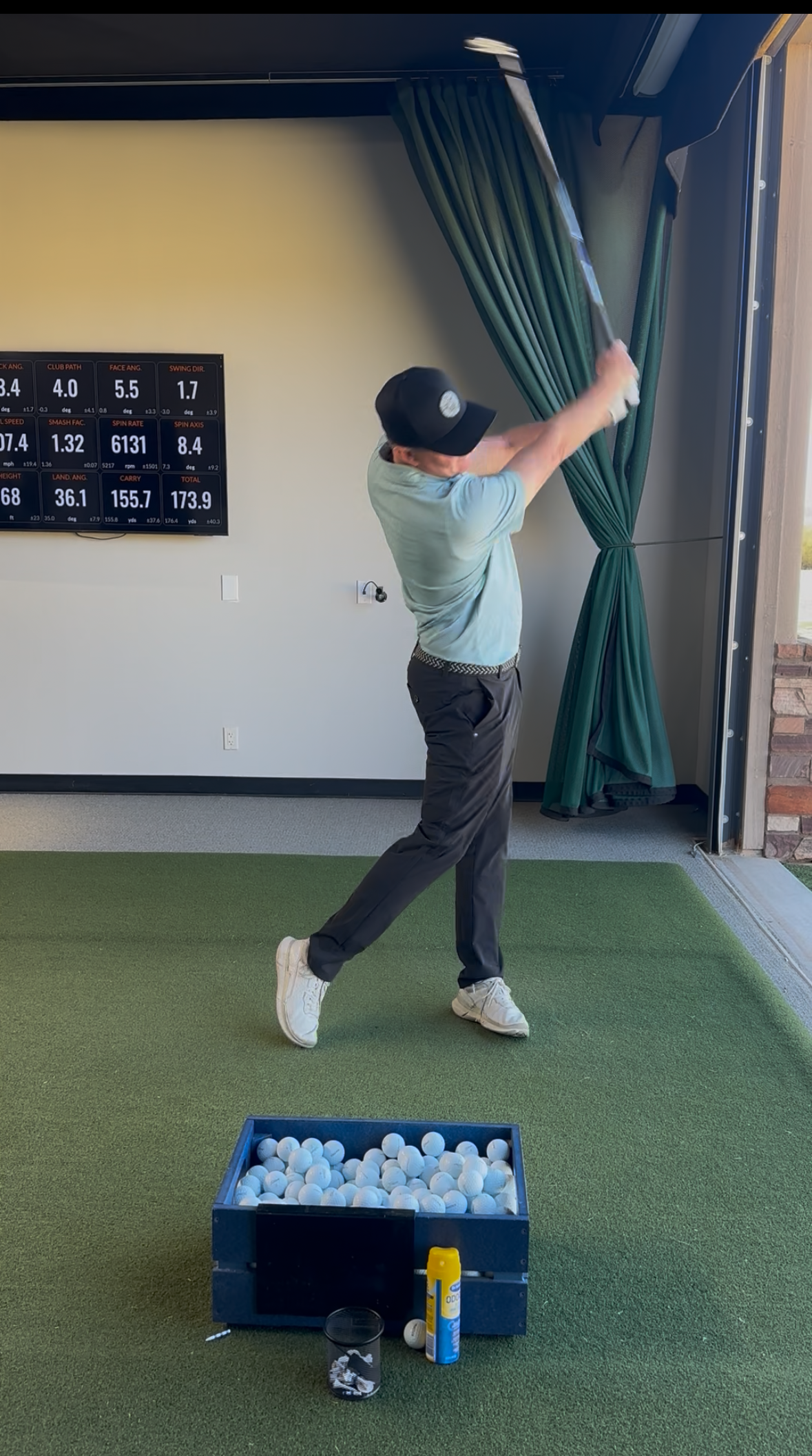 Man swinging a golf club at a driving range, wearing a cap and light blue shirt.