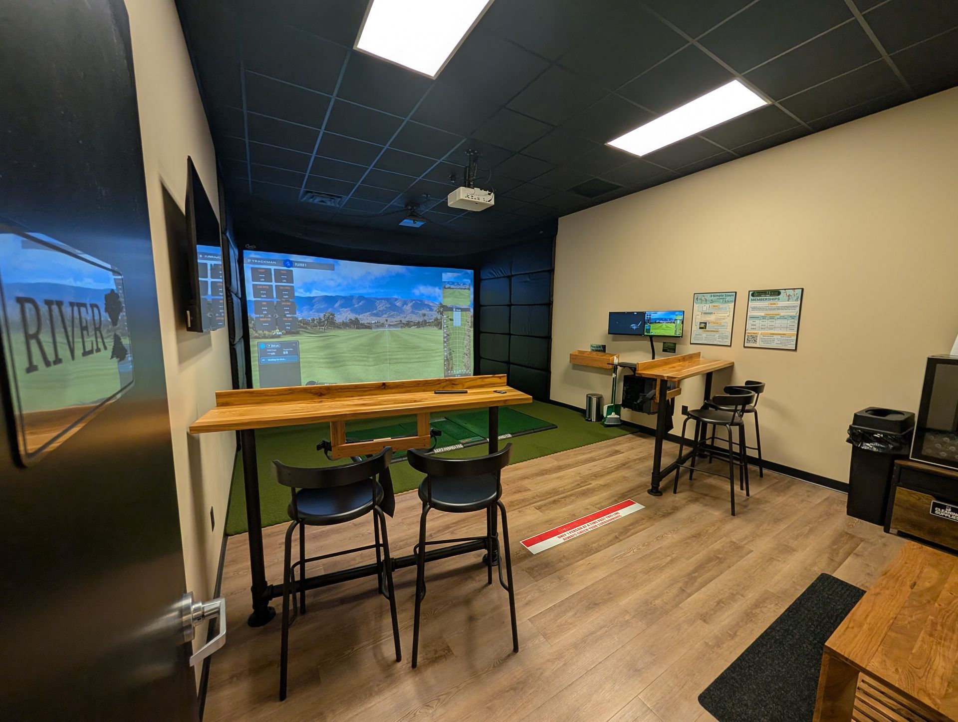 Golf simulator room: bar seating, screen displaying course, black ceiling, tan walls.