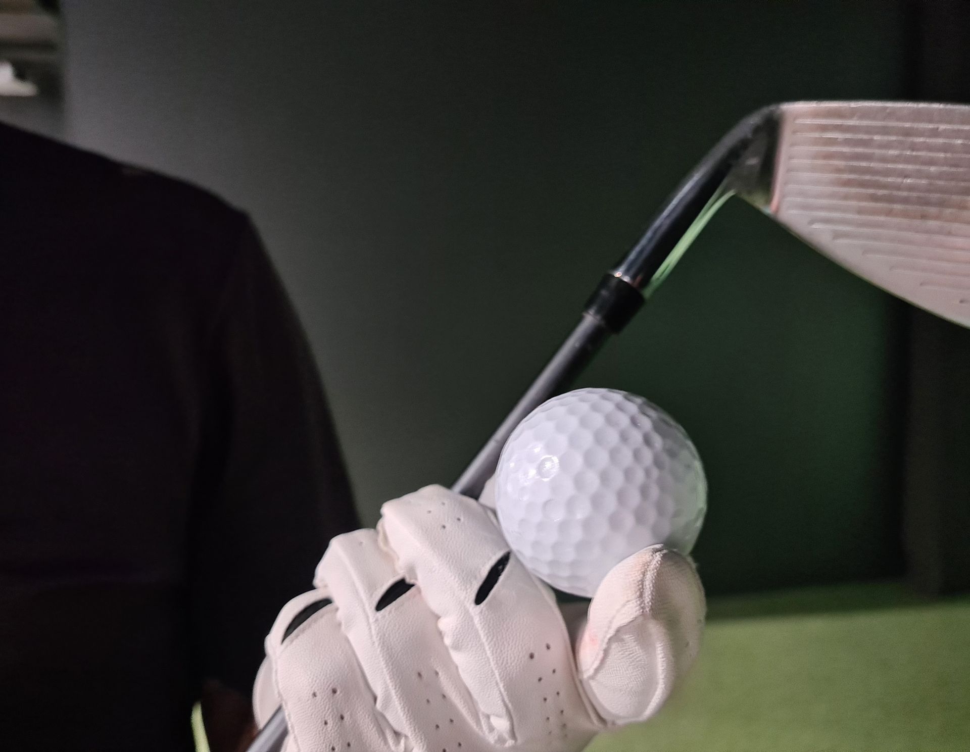 Golfer's gloved hand holds a golf ball near an iron club head; indoors, gray tones.