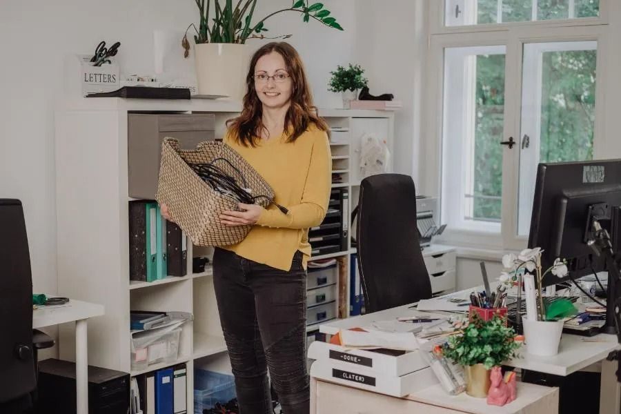 A woman with glasses in a yellow sweater holds a basket of wires in an office setting.