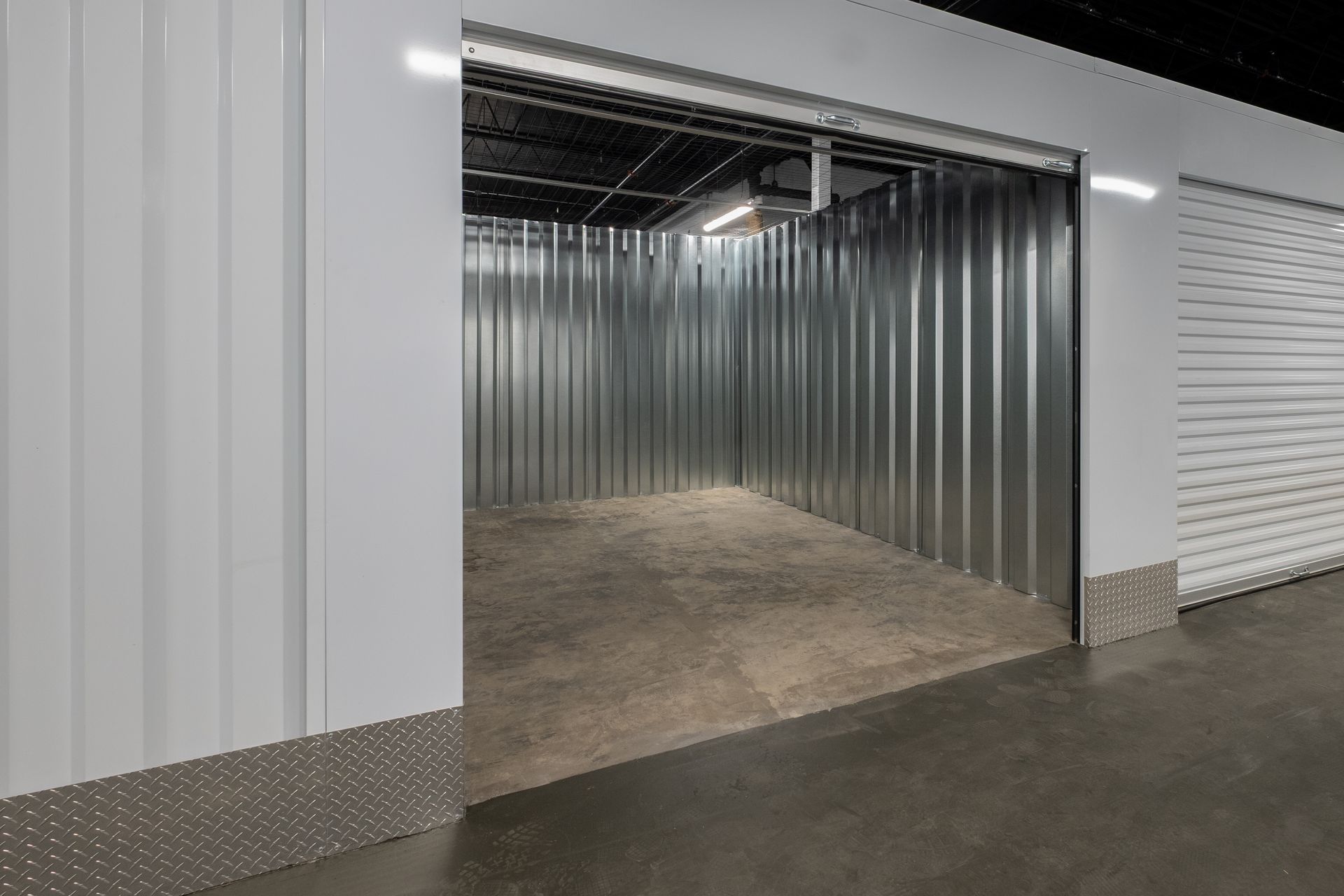Empty indoor self storage unit with metal walls, concrete flooring, and an open roll up door.