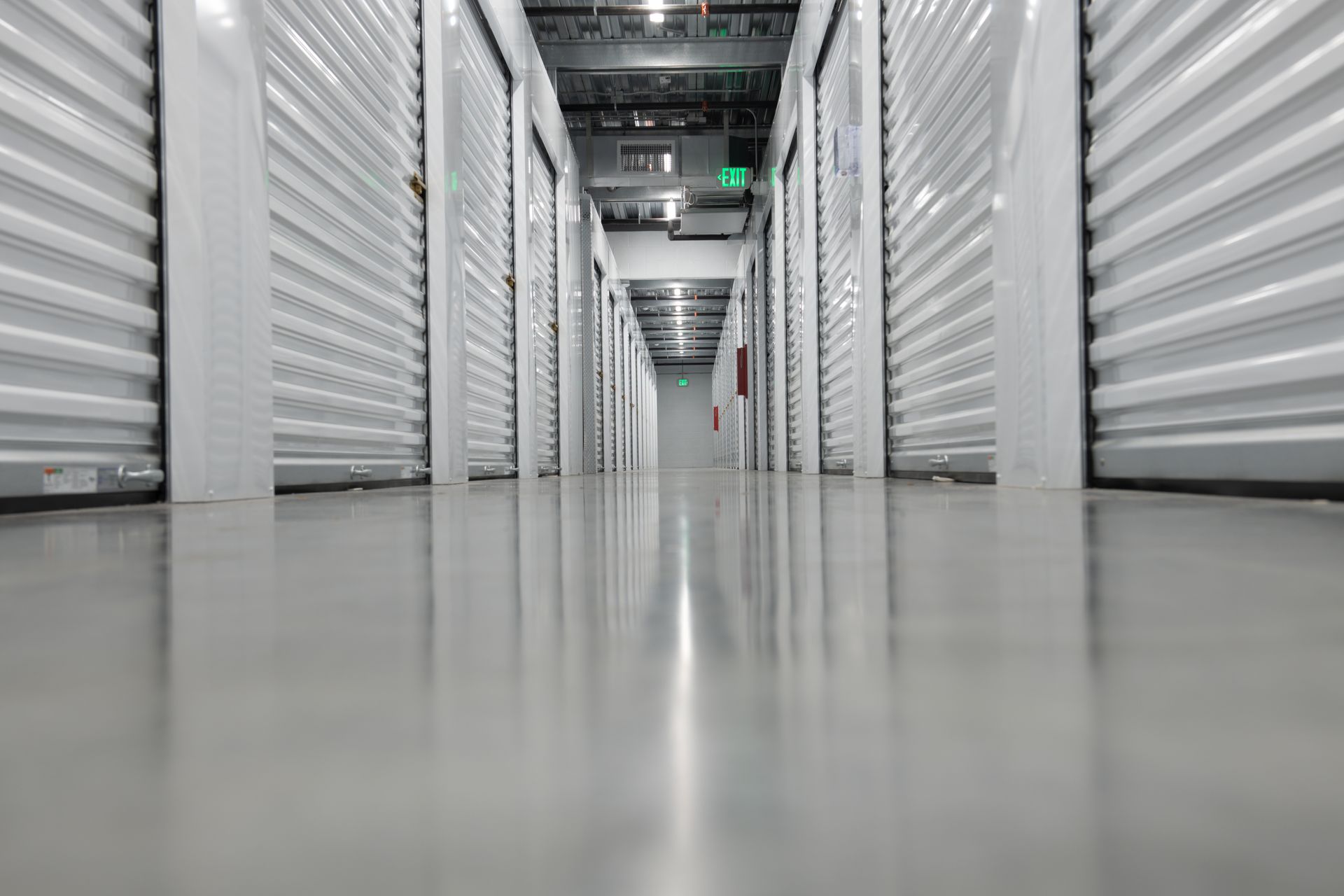 Long hallway lined with closed metal self storage unit doors and polished concrete flooring.