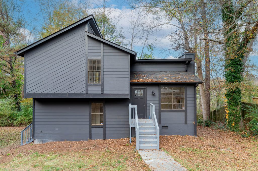 A gray house with stairs leading up to it is surrounded by trees and leaves.