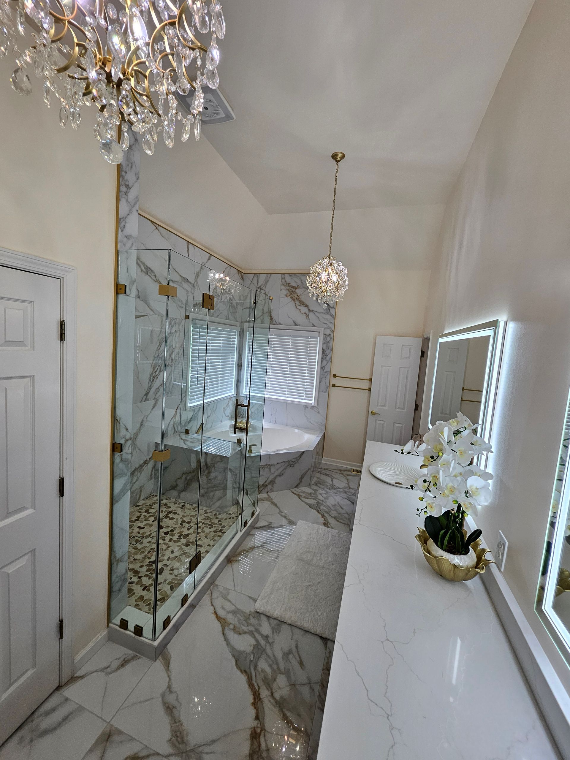 A bathroom with a walk in shower , sinks , mirrors and a chandelier.