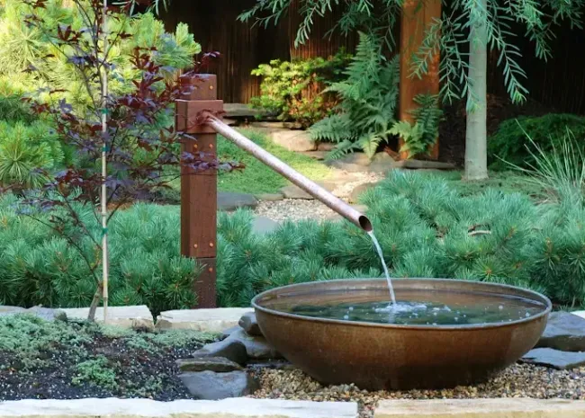 A copper pipe mounted on a wooden post pours water into a wide, rust-colored metal bowl in a lush garden setting.