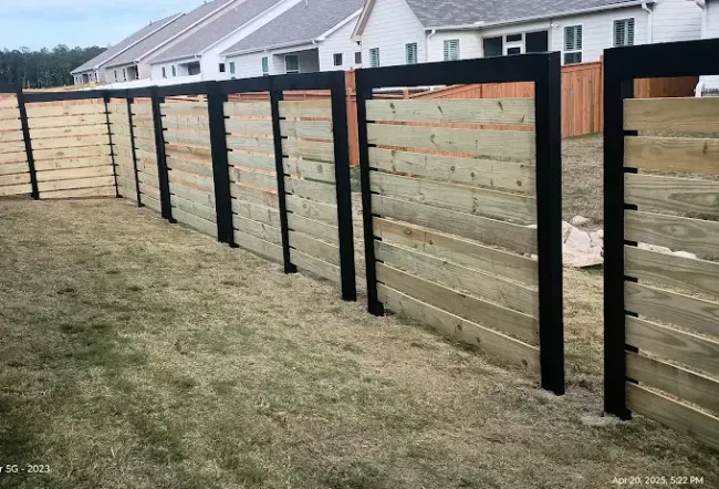 A horizontal wood-plank privacy fence featuring black metal support frames, bordering a grassy yard with houses in the back.