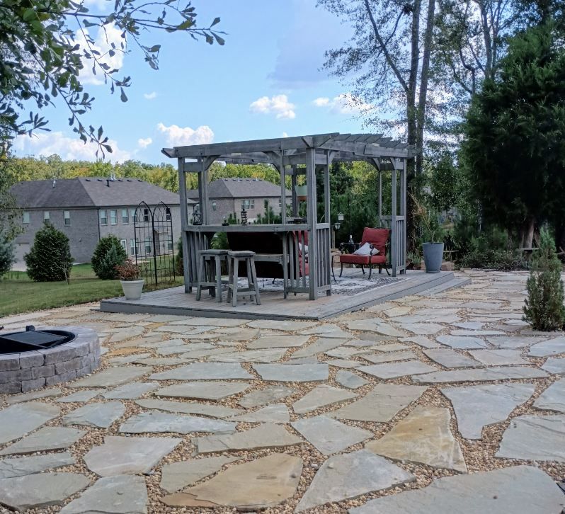 A stone patio with a gray pergola, a bar table with two stools, and a fire pit in a sunny backyard setting.