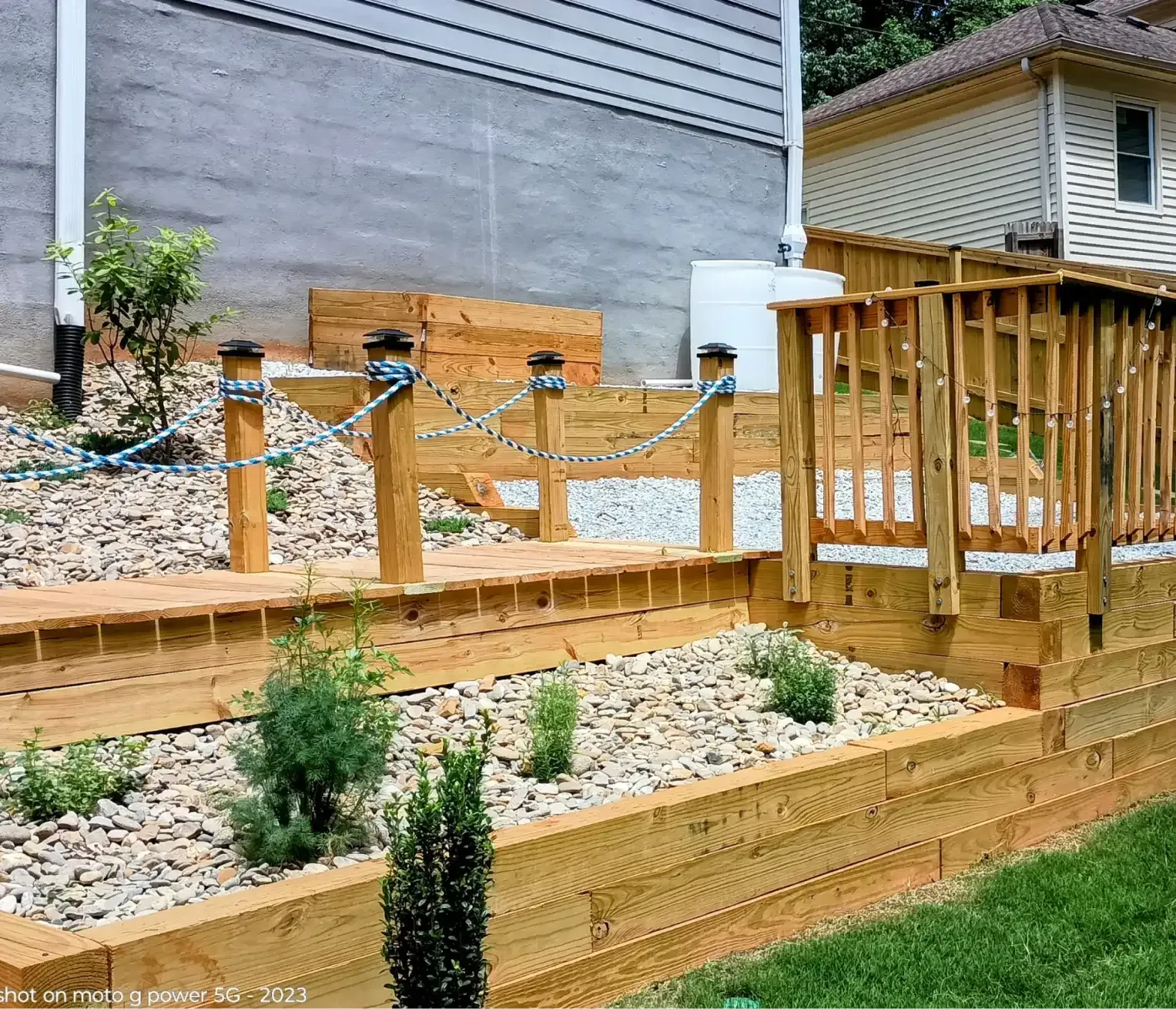 Tiered wooden garden beds with gravel, small plants, and a wooden rope-railed path next to a house exterior.
