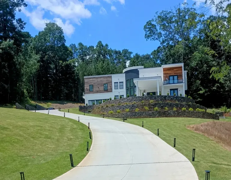 A modern, multi-story house sits atop a stone retaining wall at the end of a long, curved concrete driveway.