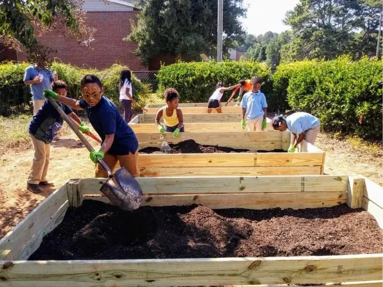 Students work together, using shovels to fill wooden garden beds with soil outdoors.