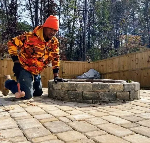 A person in an orange hoodie and hat kneels on a stone patio while building a circular stone fire pit in a backyard.