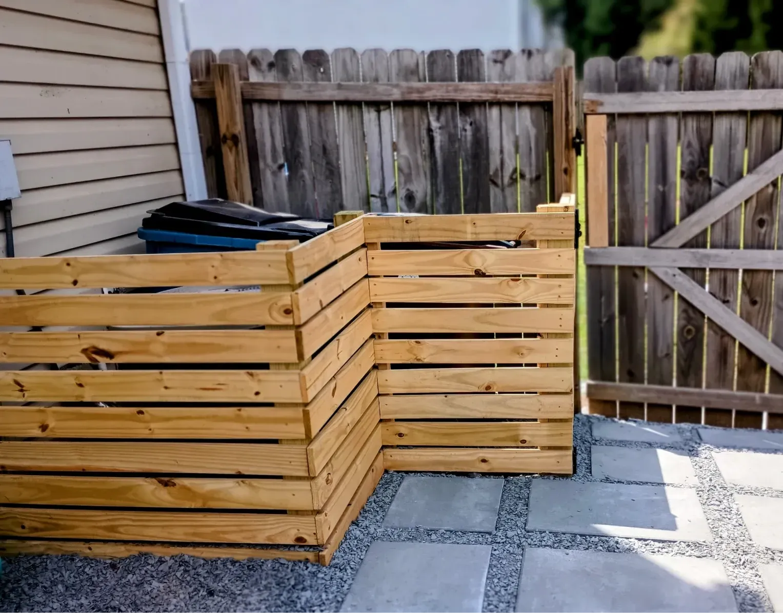 A wooden slat privacy screen conceals trash cans on a gravel patio next to a house and a wooden fence.