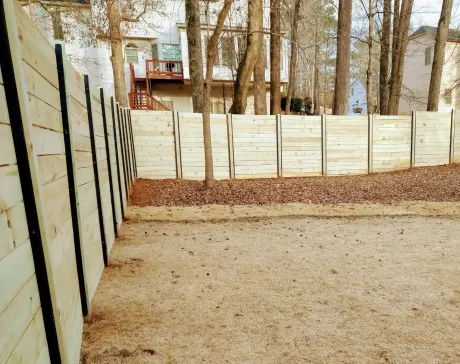 A horizontal wooden fence with black metal posts stands in a backyard with dry grass and autumn leaves.