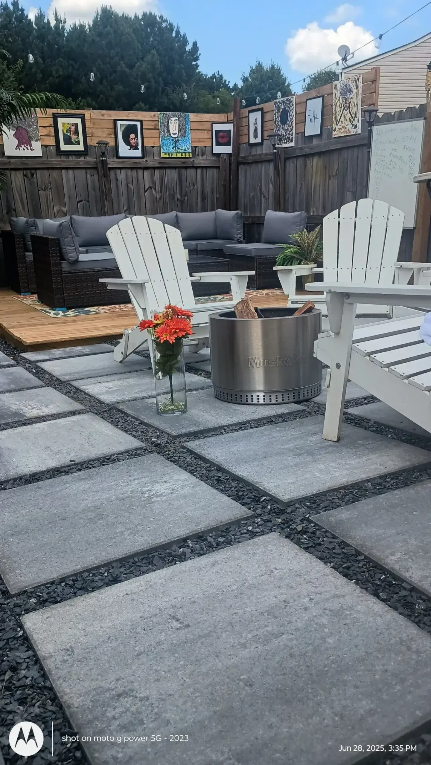 A modern backyard patio features large square concrete pavers, two white Adirondack chairs, a metal fire pit, and a sofa.