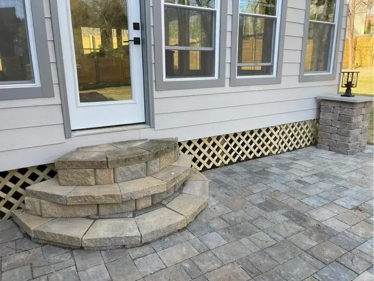 Light-colored house siding with a white door, stone steps, and lattice skirting above a stone paver patio.
