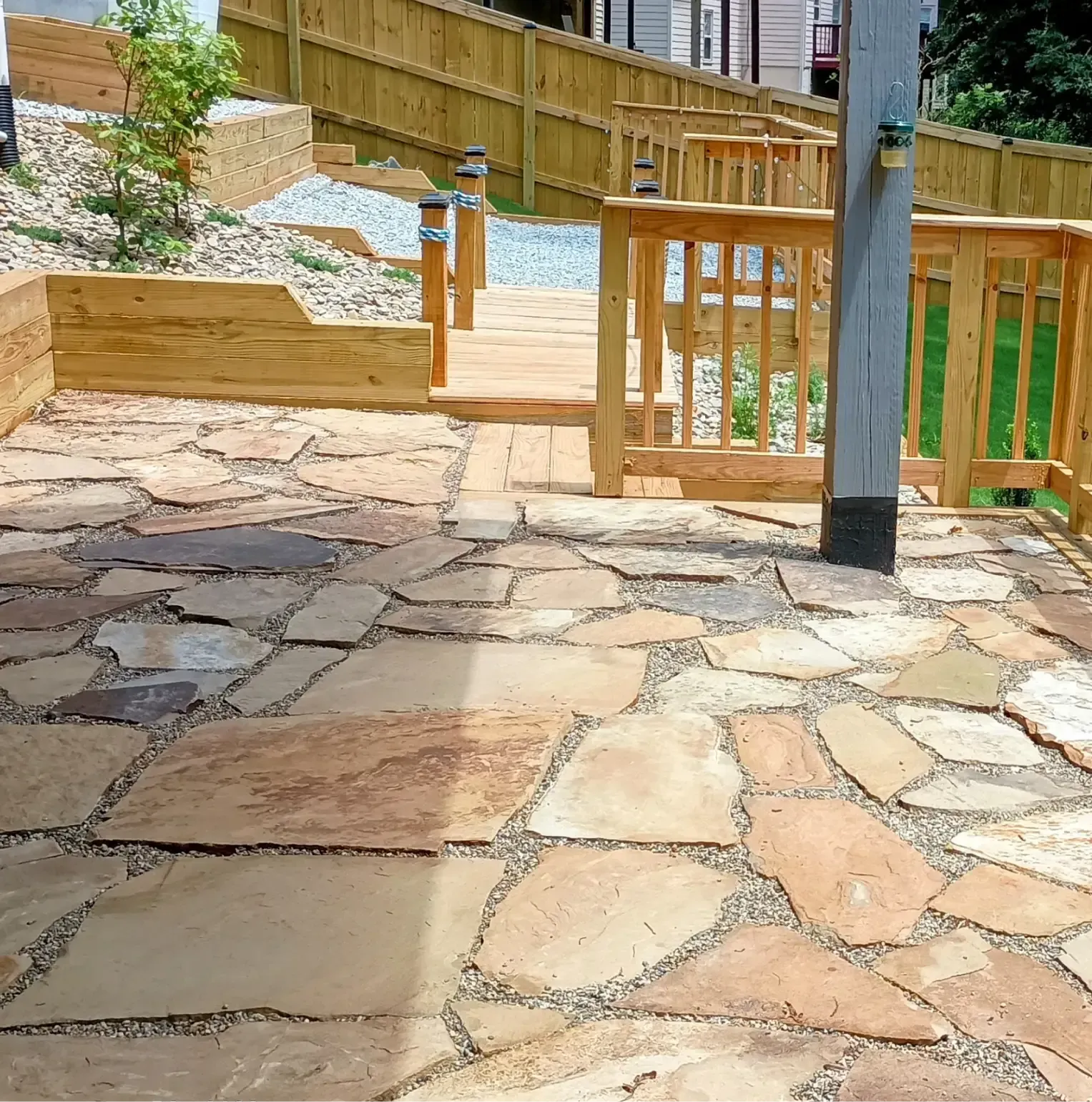 A patio made of irregular natural stone slabs with gravel joints, leading to a wooden deck and fence in a backyard.