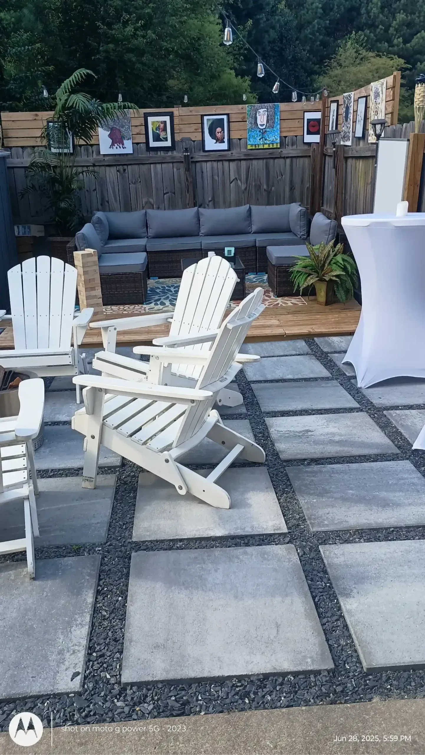 A backyard patio with white Adirondack chairs on concrete pavers, a gray sectional sofa, and a draped cocktail table.