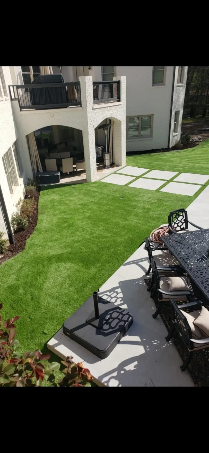 A high-angle view of a backyard with a grass lawn, paved patio, and a covered lower-level porch on a two-story home.