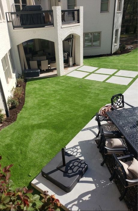 A high-angle view of a backyard featuring a lush green lawn, a stone patio with outdoor seating, and a paved walkway.