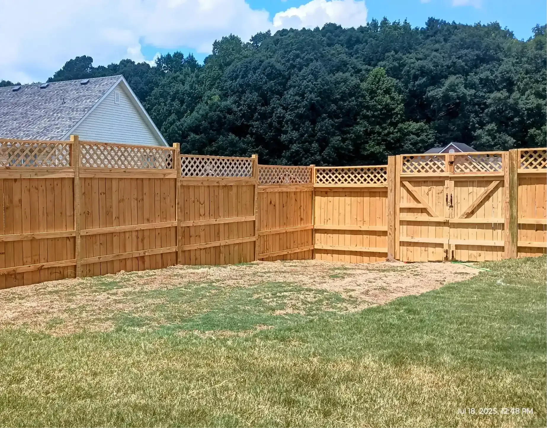 A wooden privacy fence with a decorative lattice top spans across a grassy backyard under a bright blue sky.