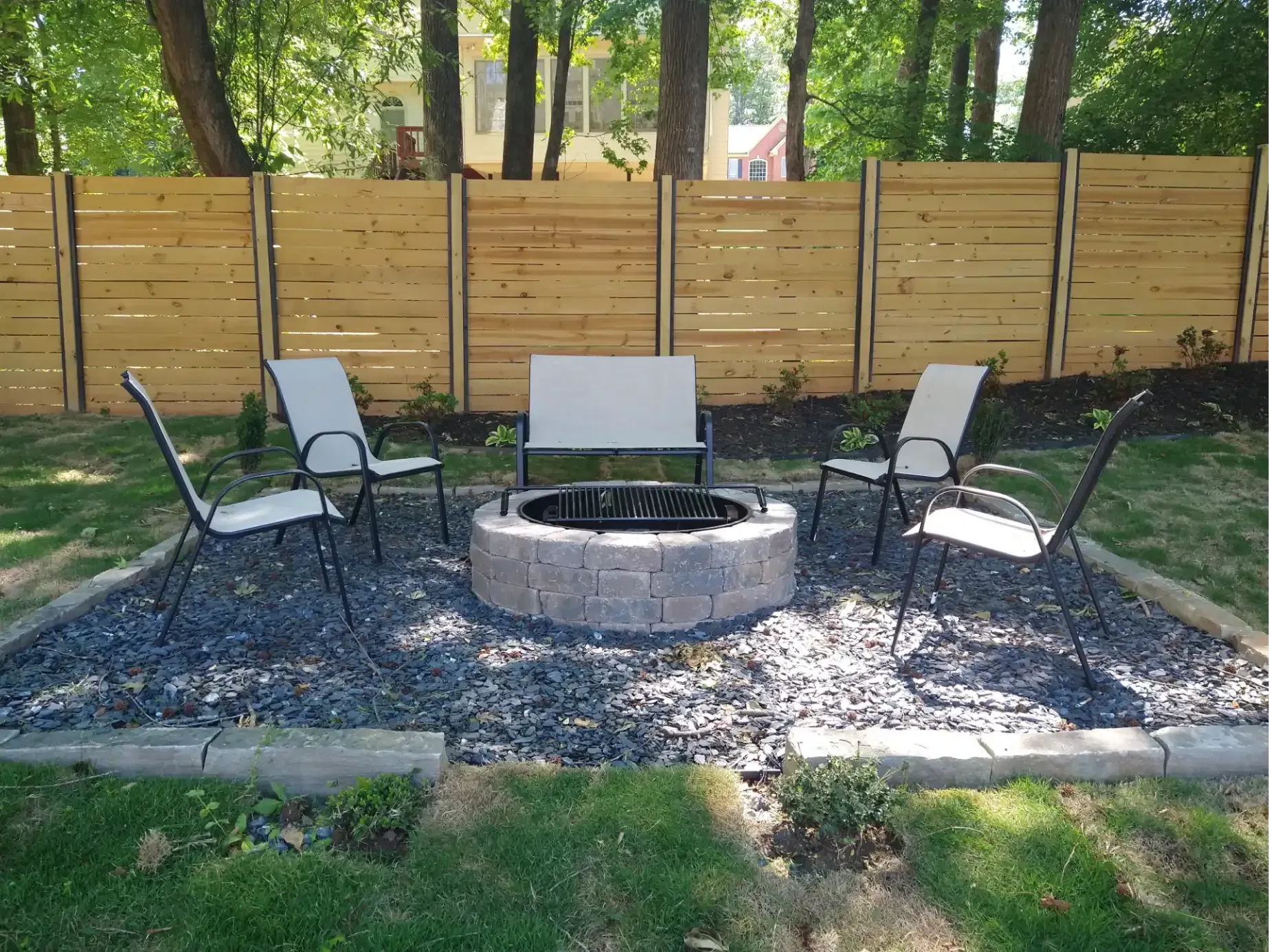 A circular stone fire pit sits in a gravel patio area with four outdoor chairs and a wooden fence in the background.