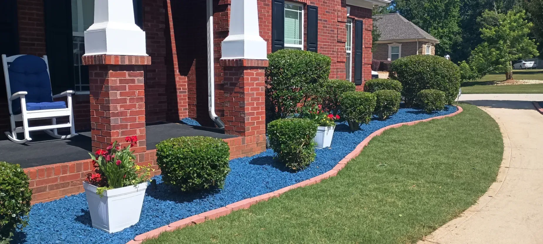 A brick home porch features green shrubs and a potted plant set against a bed of bright blue decorative landscaping rocks.