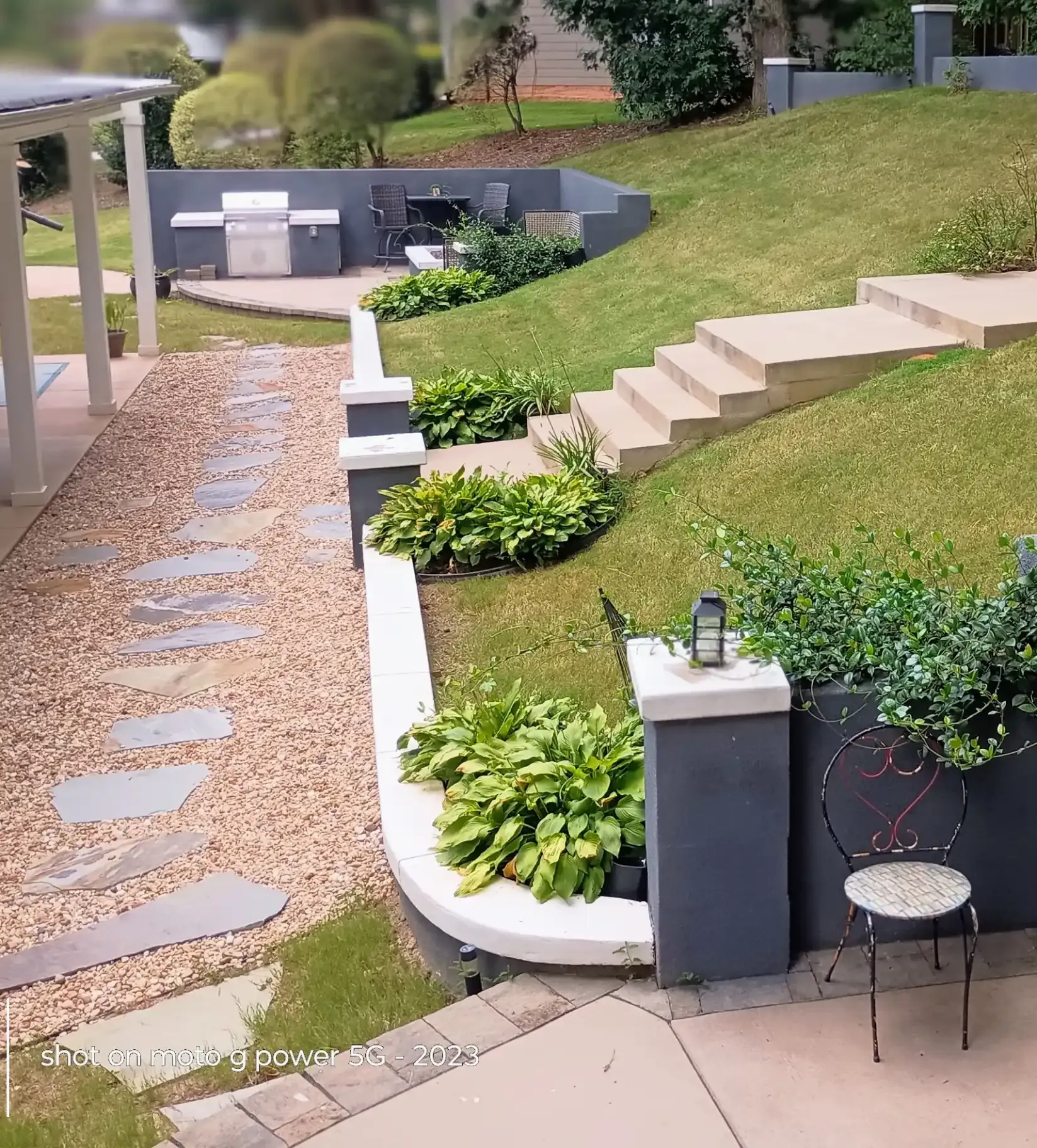 A stone-paved gravel path leads past tiered retaining walls, green plants, and concrete stairs toward a patio with a grill.