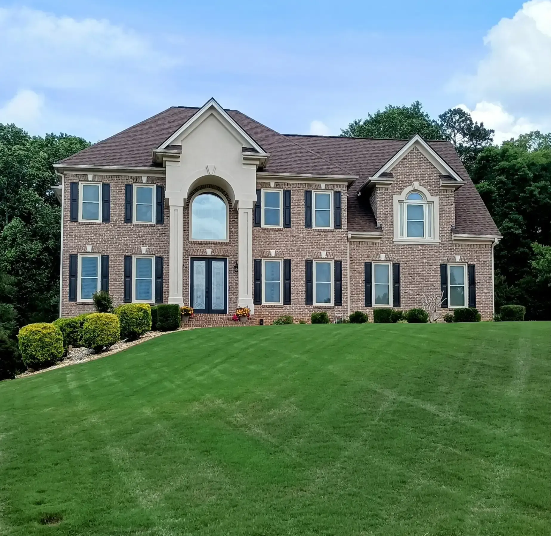 A two-story brick suburban house with a central arched entryway and dark shutters, surrounded by a large green lawn.