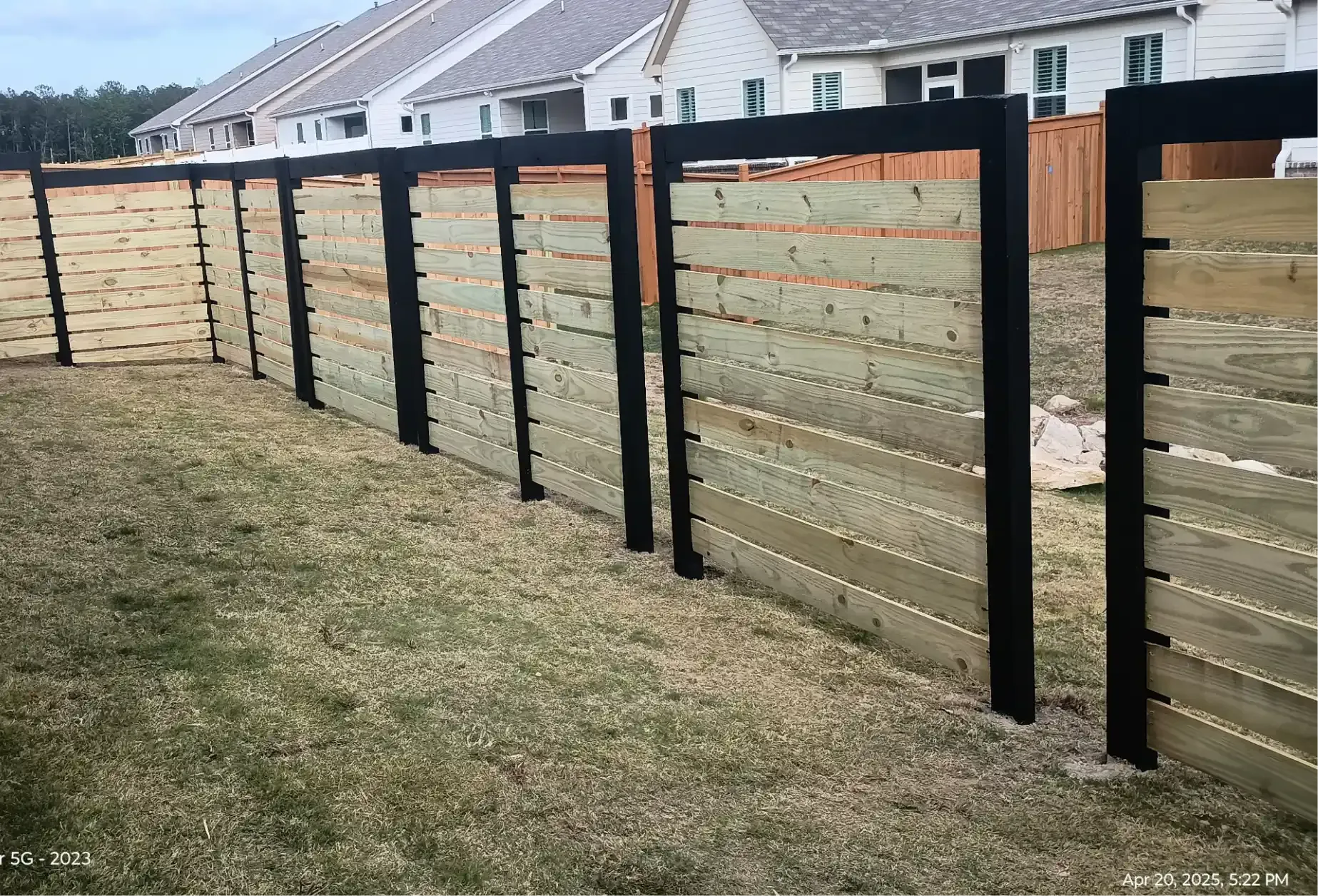 A modern horizontal wood fence with black metal posts set against suburban houses and a grassy lawn.