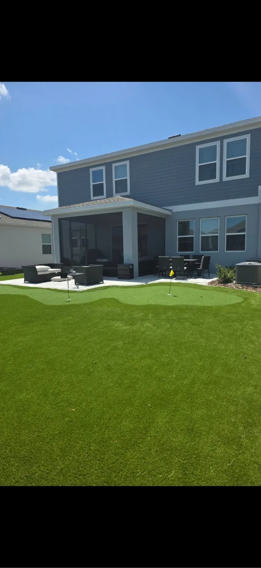 A backyard featuring a blue two-story house with a screened-in patio, a putting green, and a large lawn under a blue sky.