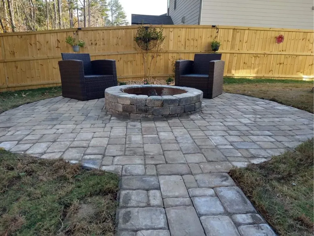 A circular stone fire pit sits on a grey paver patio in a backyard with two dark wicker chairs and a wooden fence.