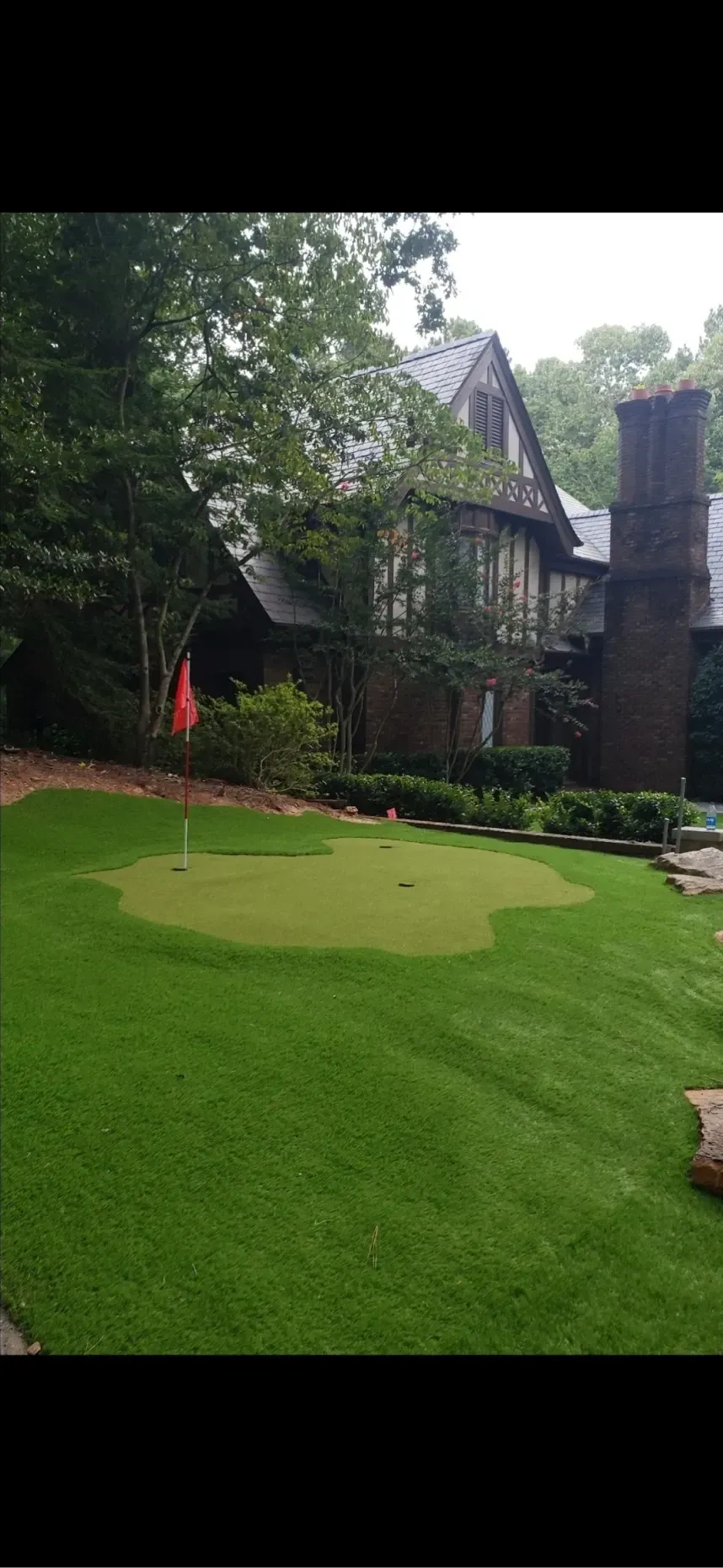 A backyard putting green with a flag pole sits in front of a large, multi-story Tudor-style home nestled in the woods.