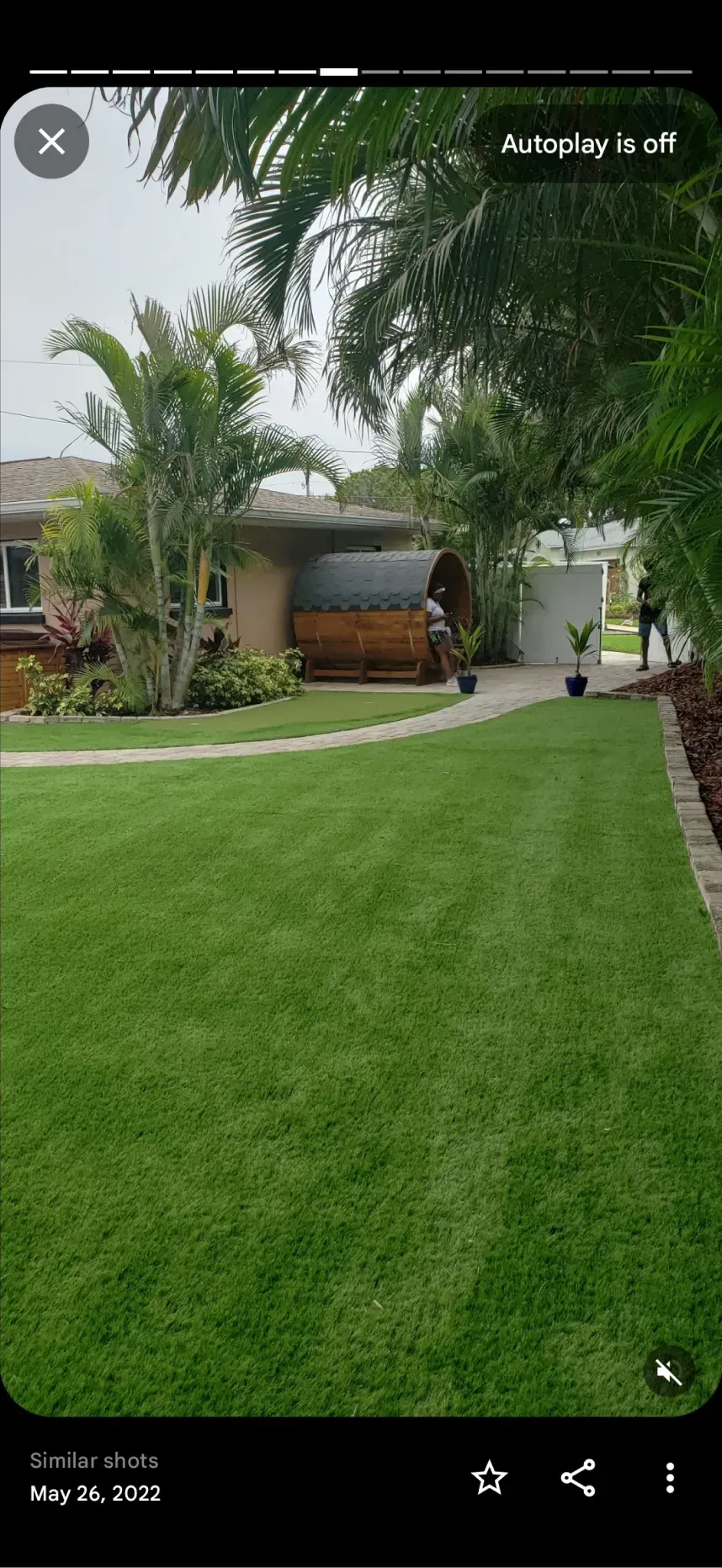 A well-manicured lawn leads to a house with a wooden barrel sauna in the backyard on a cloudy day.