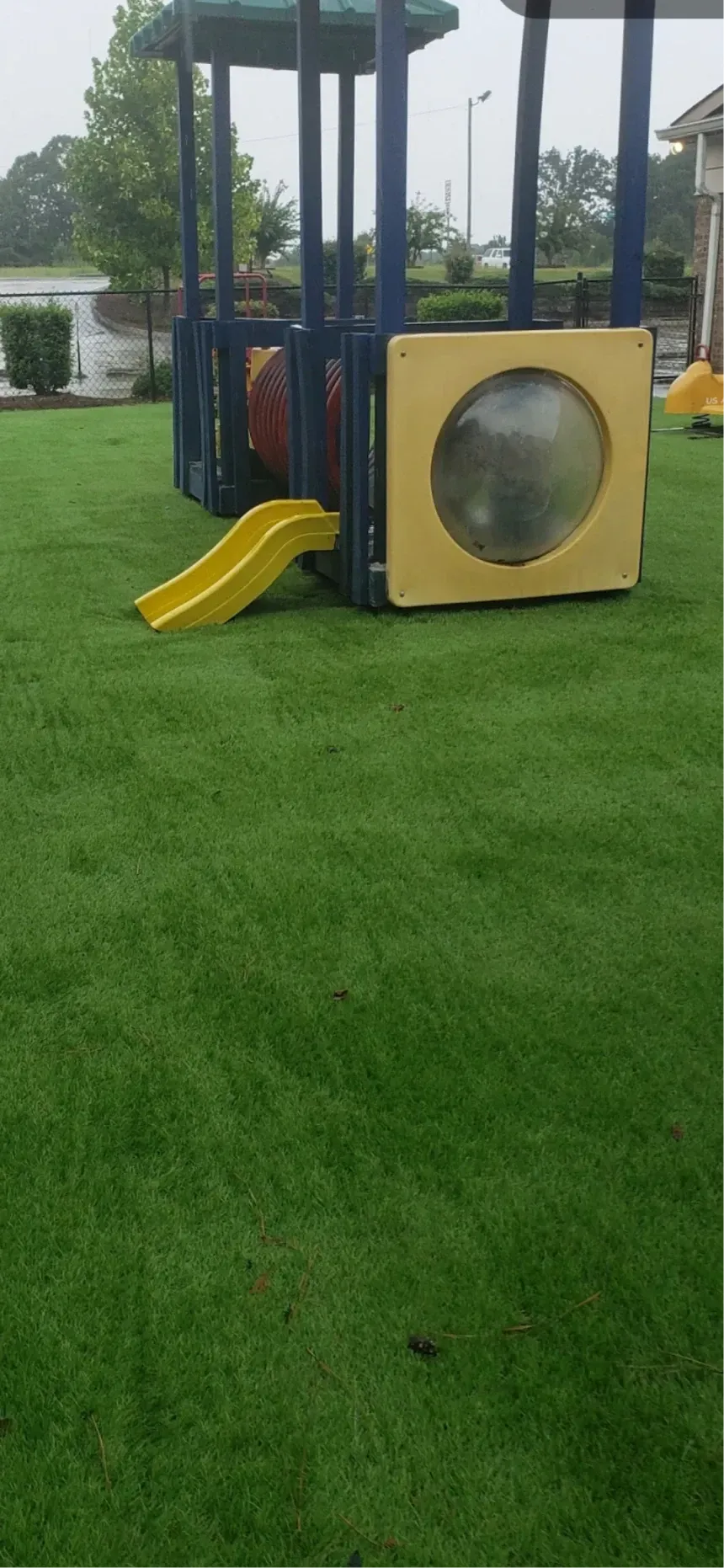 A playground structure with a yellow slide and tunnel sits in the middle of a vibrant green grassy lawn on a rainy day.