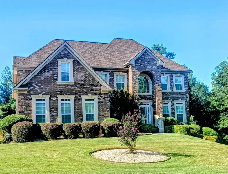 A large, two-story brick house with a dark shingled roof, set behind a manicured lawn with shrubs and a circular garden bed.