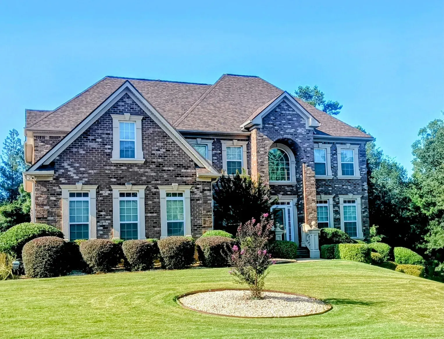 A large two-story brick suburban home with a dark shingled roof, set behind a green lawn with a small, circular garden.