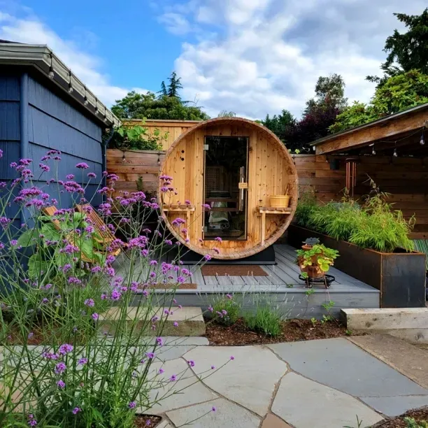 A round wooden barrel sauna sits on a backyard deck, surrounded by plants and a flagstone path, next to a blue building.