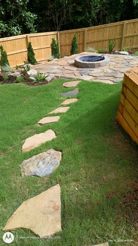 A stone pathway leads across a green lawn to a circular stone fire pit in a backyard with a wooden fence.