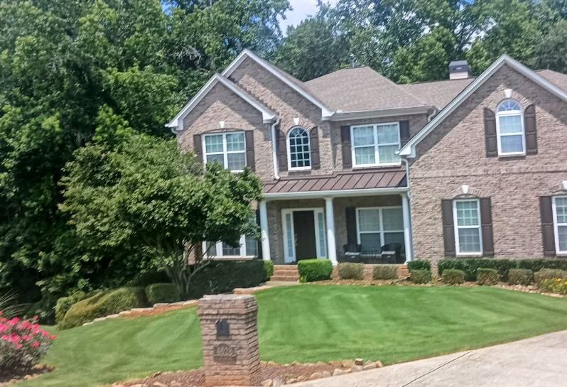 A large, two-story brick house with a green lawn, dark shutters, and a metal roof porch, set against a backdrop of trees.