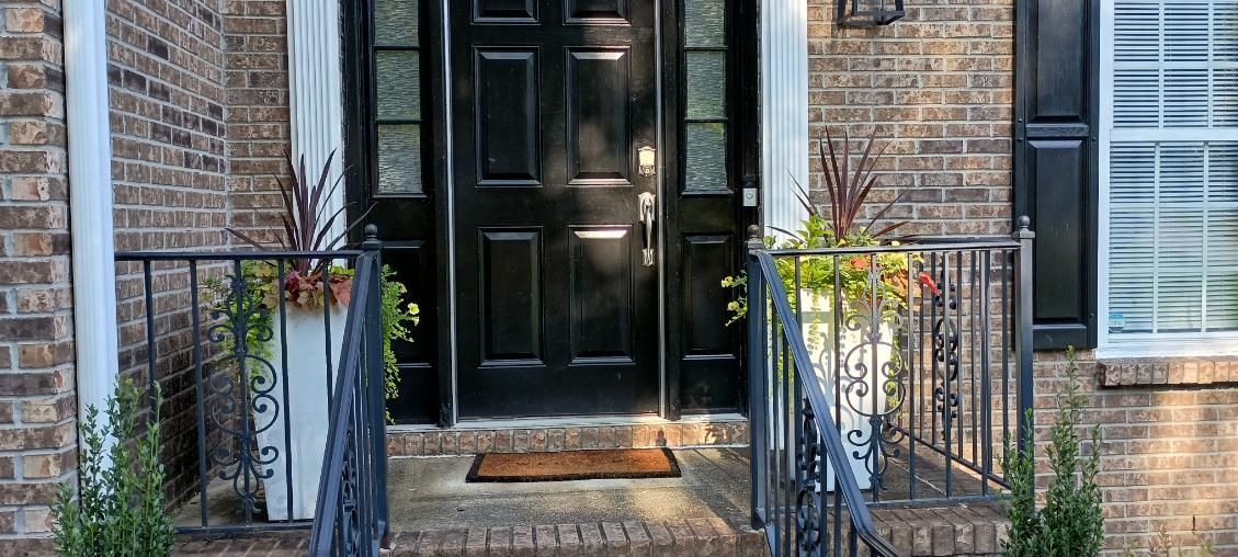 A brick home entryway featuring a black front door with side windows, iron railings, and two potted plants on the steps.