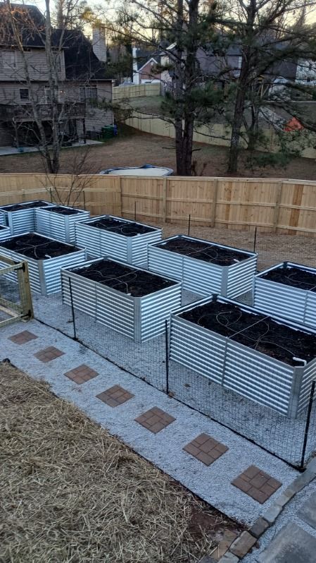 A series of raised, corrugated metal garden beds filled with dark soil on a gravel path in a fenced backyard.