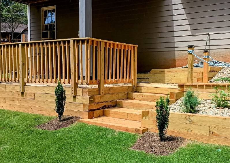 A wooden deck with a railing and steps leading to a house, featuring two small evergreen trees planted in mulch beds.