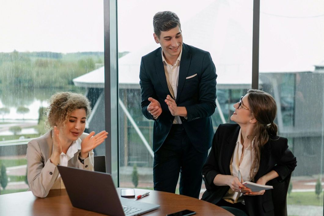 Three coworkers in a bright conference room discussing work around a laptop and phone.